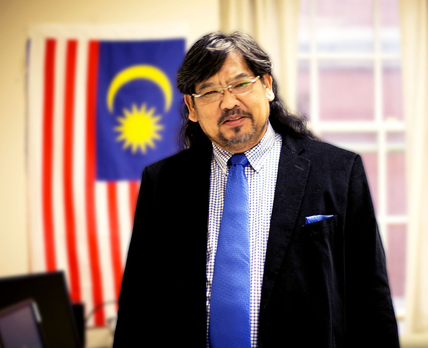 Jayum Anak Jawan standing and posing for a photo in his office with the flag of Malaysian flag in the background