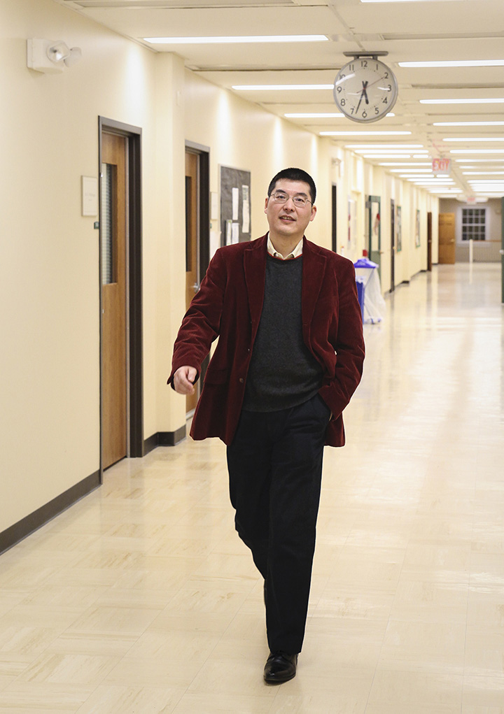 Zijian Diao walks down a hallways with a smile. He wears black slacks, a dark grey sweater, and a deep red sweater. 