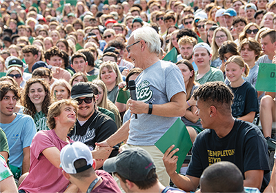 President Sherman standing in a crowd of students inside of Peden Stadium. He is holding a microphone while interacting with a boy