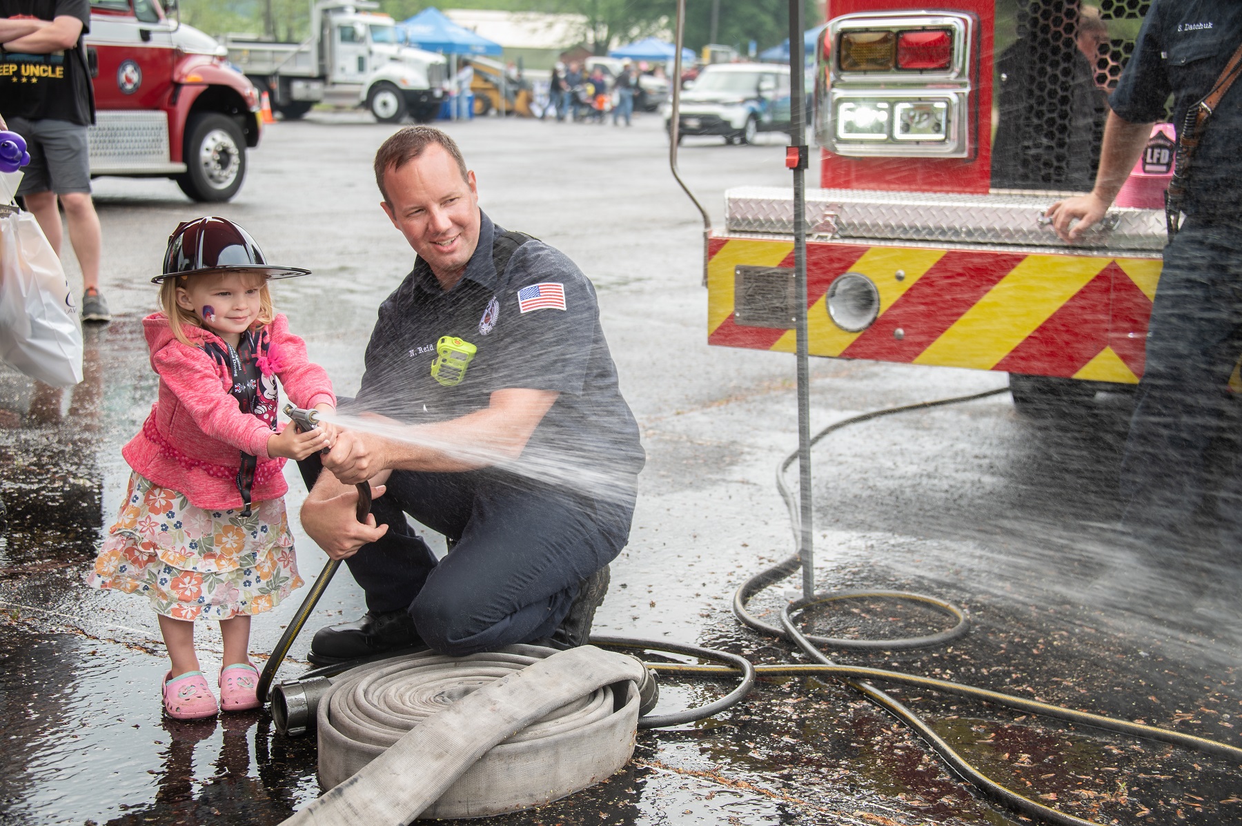 A child sprays water out of a hose while wearing a firefighters hat, and a firefighter is helping her