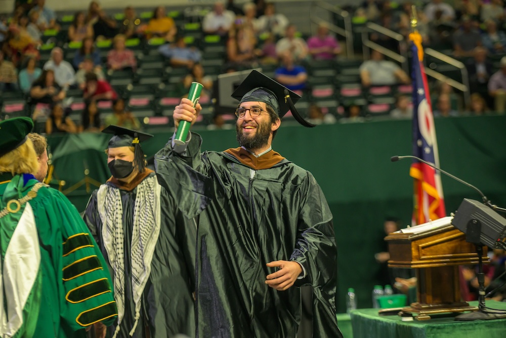 A man in academic regalia walks across the stage at Commencement holding a green tube aloft to show someone in the audience