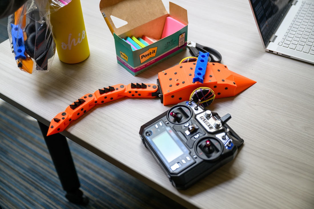 An orange and blue robot with a long tail sits beside a controller