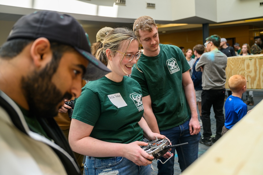 A student in a green t-shirt smiles as she holds a remote control, and two other people watch what she's looking at