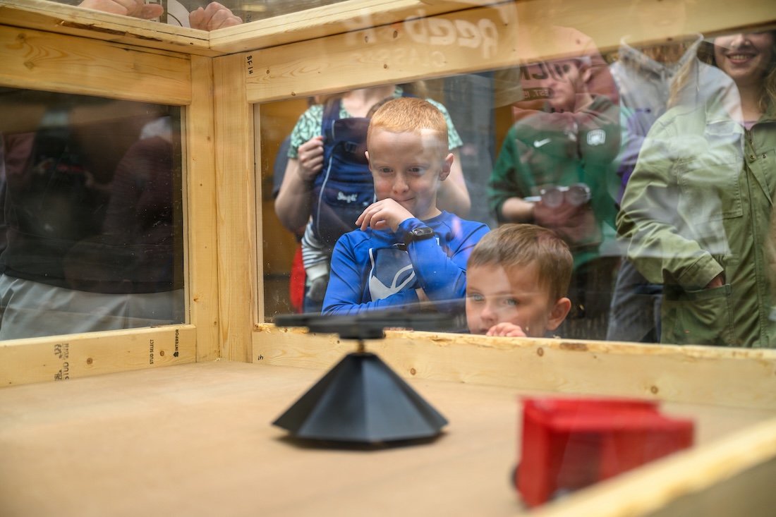 Two small robots are pictured in a plywood box with windows as children watch