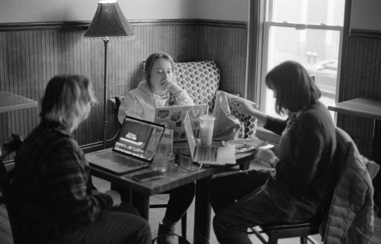 In a black and white photo, three students are seated around a table, working intently on laptops