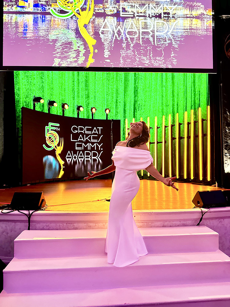 a woman in a formal gown poses exuberantly in front of a colorful stage at the Great Lakes Emmy Awards