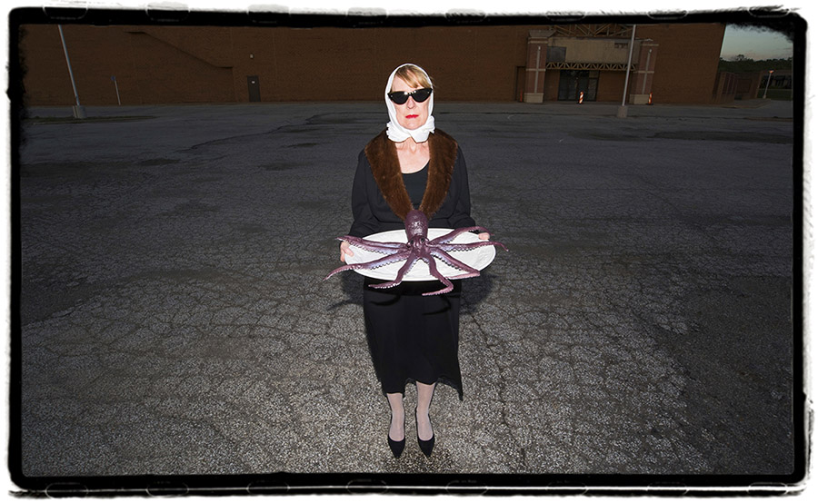 a photo of a white woman at dusk wearing sunglasses, a white kerchief covering her blonde hair, a black coat with brown fur lining and black heels; she is holding a large white plate with a purple plastic octopus on it