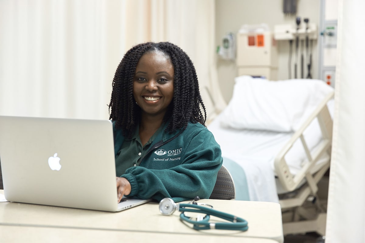 A woman in a nurse's uniform sits at a desk containing a laptop and a stethoscope in a hospital room