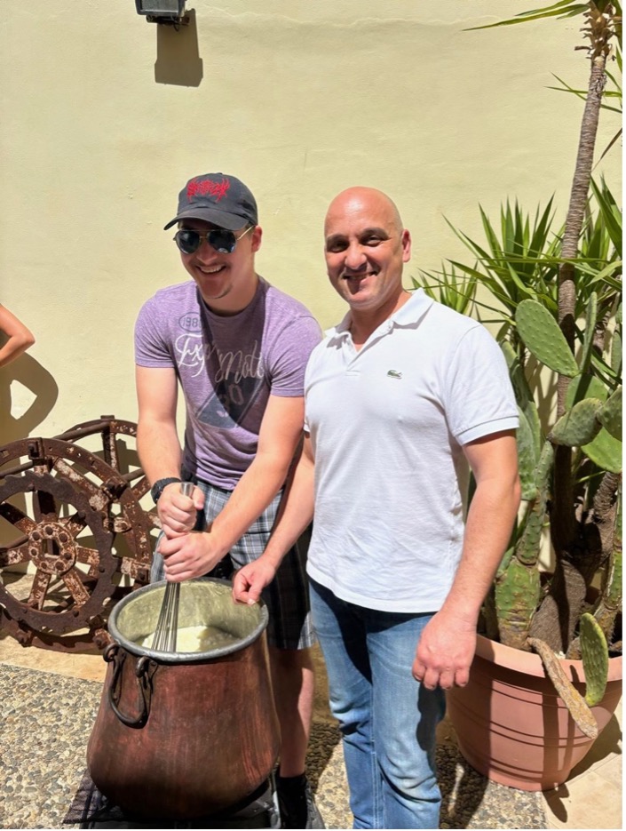 Two men pose in front of a cooking pot while one of them stirs