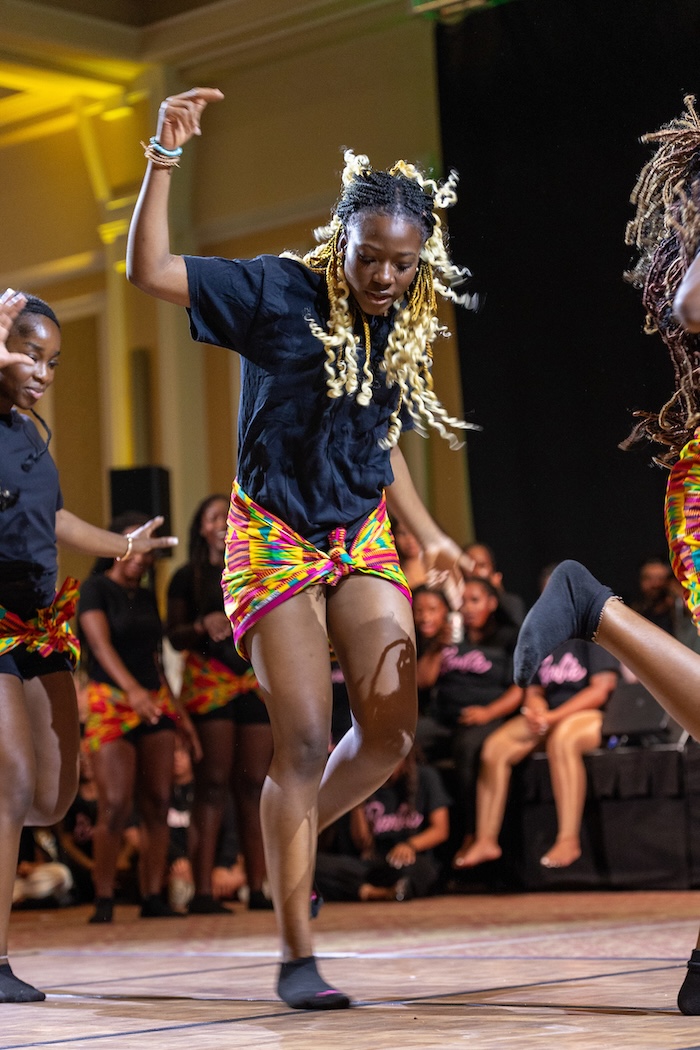 A student in a black top and colorful skirt dances with a group