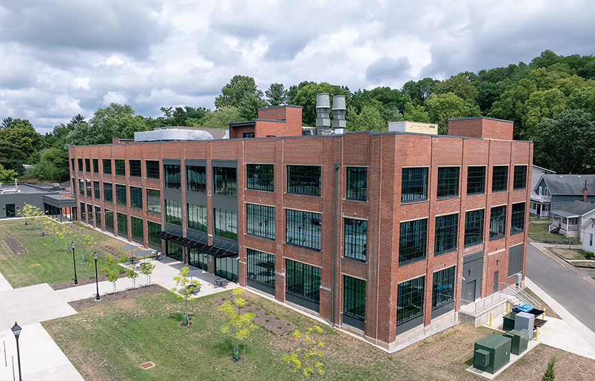 an aerial view of a three-story brick building’s exterior, which has windows covering both visible walls and is surrounded by newly planted grass and trees