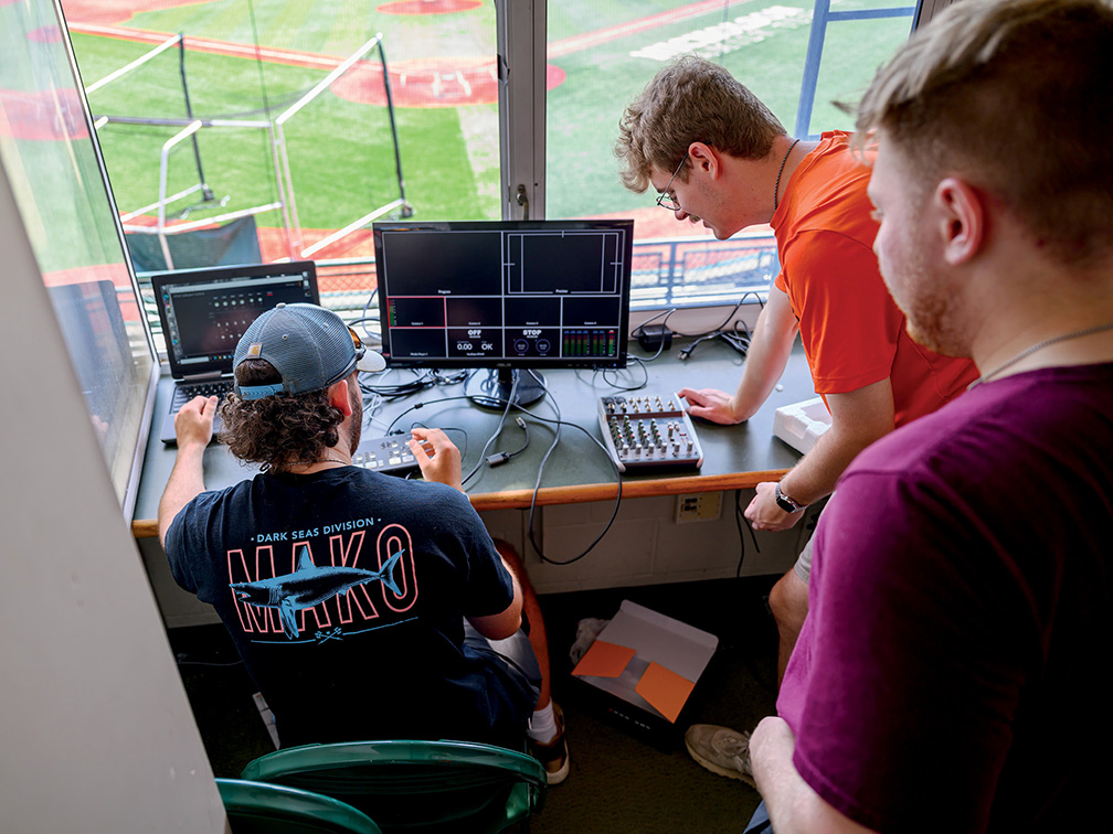 three male students look over computer equipment used to broadcast Copperheads games in the stadium's control room