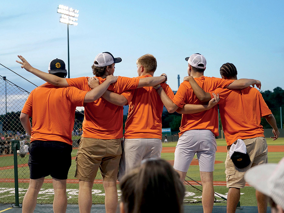 five male students in orange shirts stand with their backs to the camera, arms around each others' shoulders, watching a Copperheads game