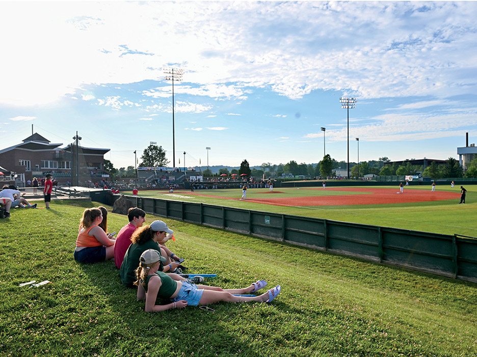 five game attendees lounge on the grass near the outfield at Bob Wren Stadium, watching a Copperheads game