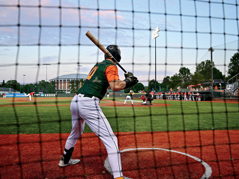 a Copperheads baseball player holds the bat as he waits on deck, viewed from behind a black safety net