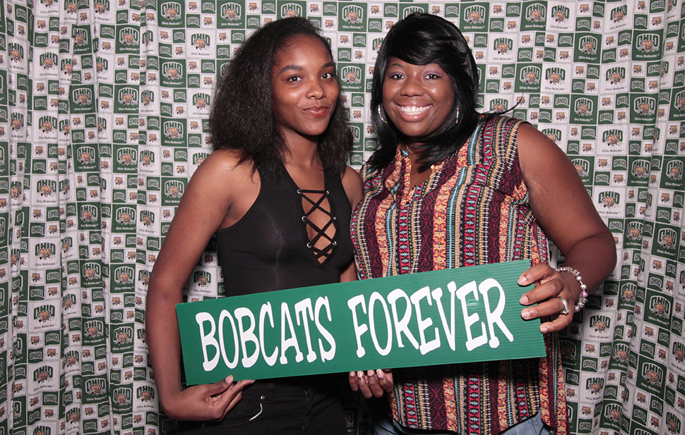 two Black women pose with a “Bobcats forever” sign in front of an Ohio University-themed photo booth backdrop