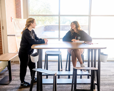 Two students converse, one seated at a tall table and the other standing