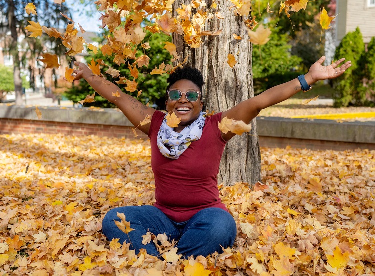 An OHIO student sits in yellow leaves  on the ground on OHIO's Athens campus and throws leaves into the  air