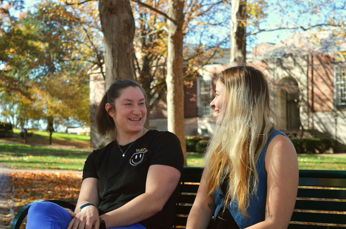 Two women sit on a bench outside, one is smiling at the other