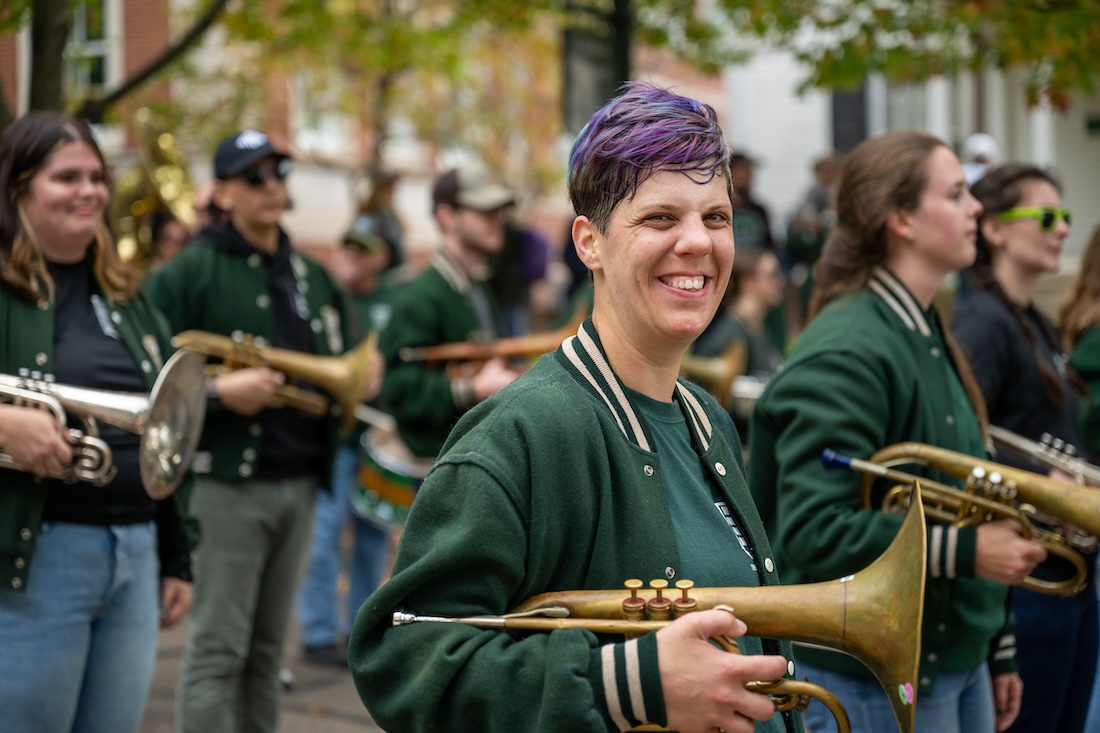 Standing in a parade with other alumni band members, a person with purple hair holding a trumpet smiles at the camera