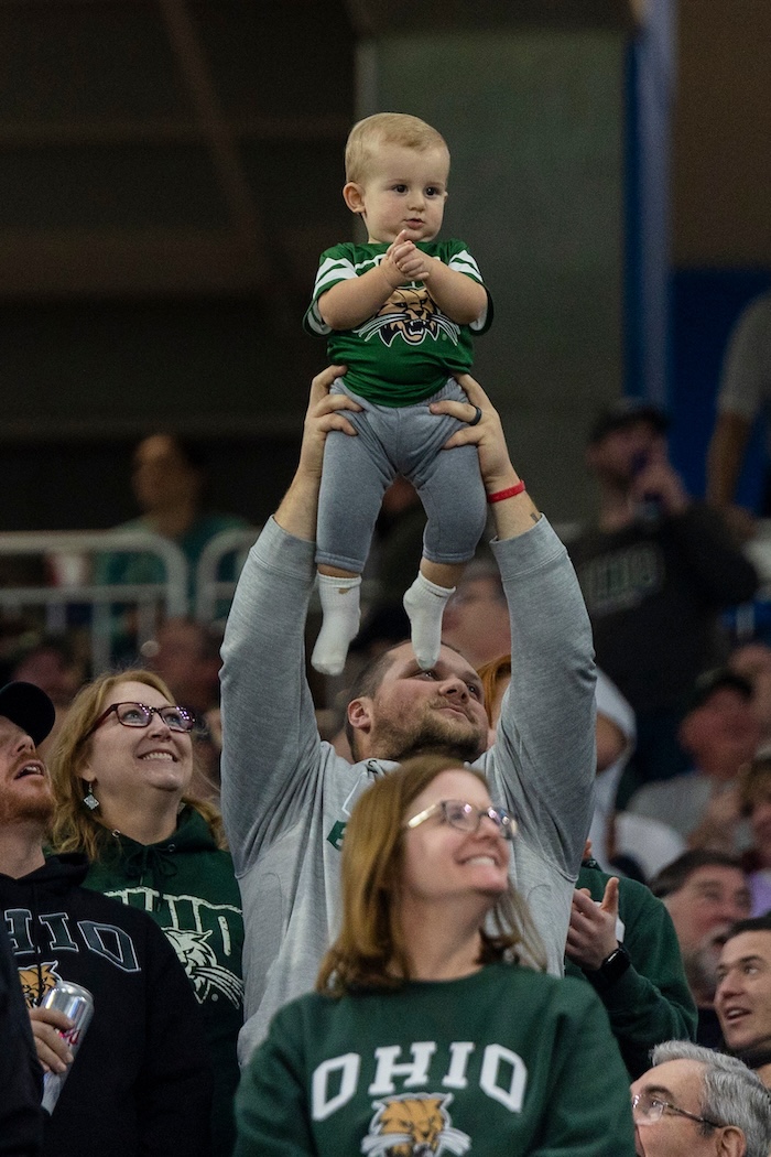 A baby in Bobcat green is held above the crowd at a football game