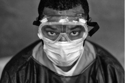 A medical professional wearing full protective gear gazes directly at the camera. Black and white image