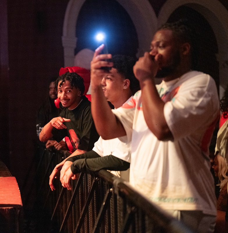 Audience members take photos and one individual smiles at the camera at the Mariah The Scientist performance