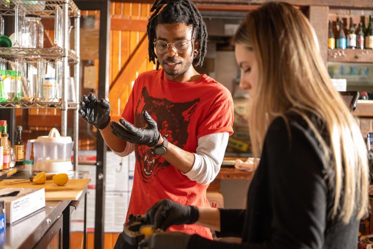 Two people stand in a laboratory, one pouring a substance while the other looks on, gesturing instructions