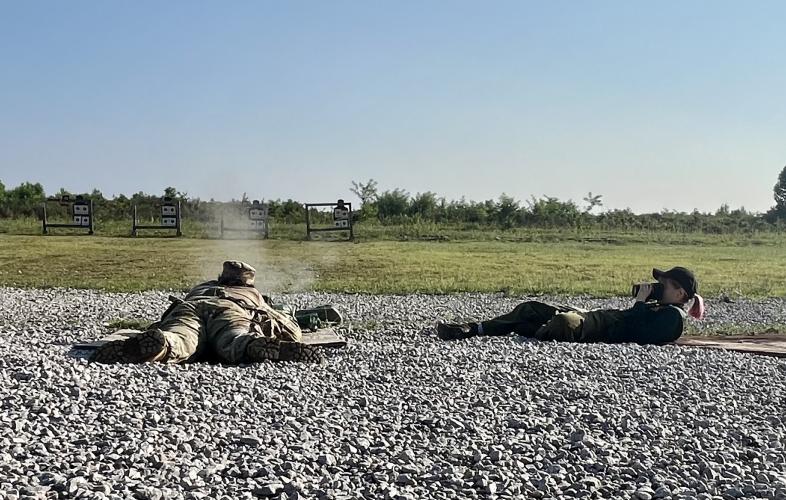 On a gravel surface, a soldier lying on the ground shoots at a target in the near distance while a photographer lies beside him, taking a picture