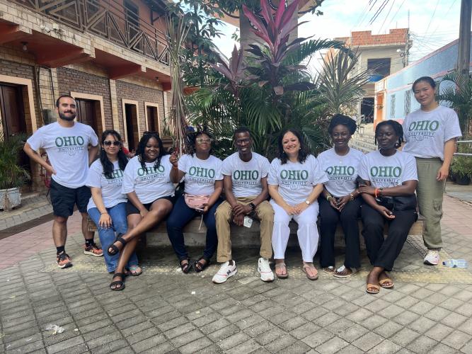 A group of students wearing OHIO t-shirts are seated under a tree, smiling at the camera