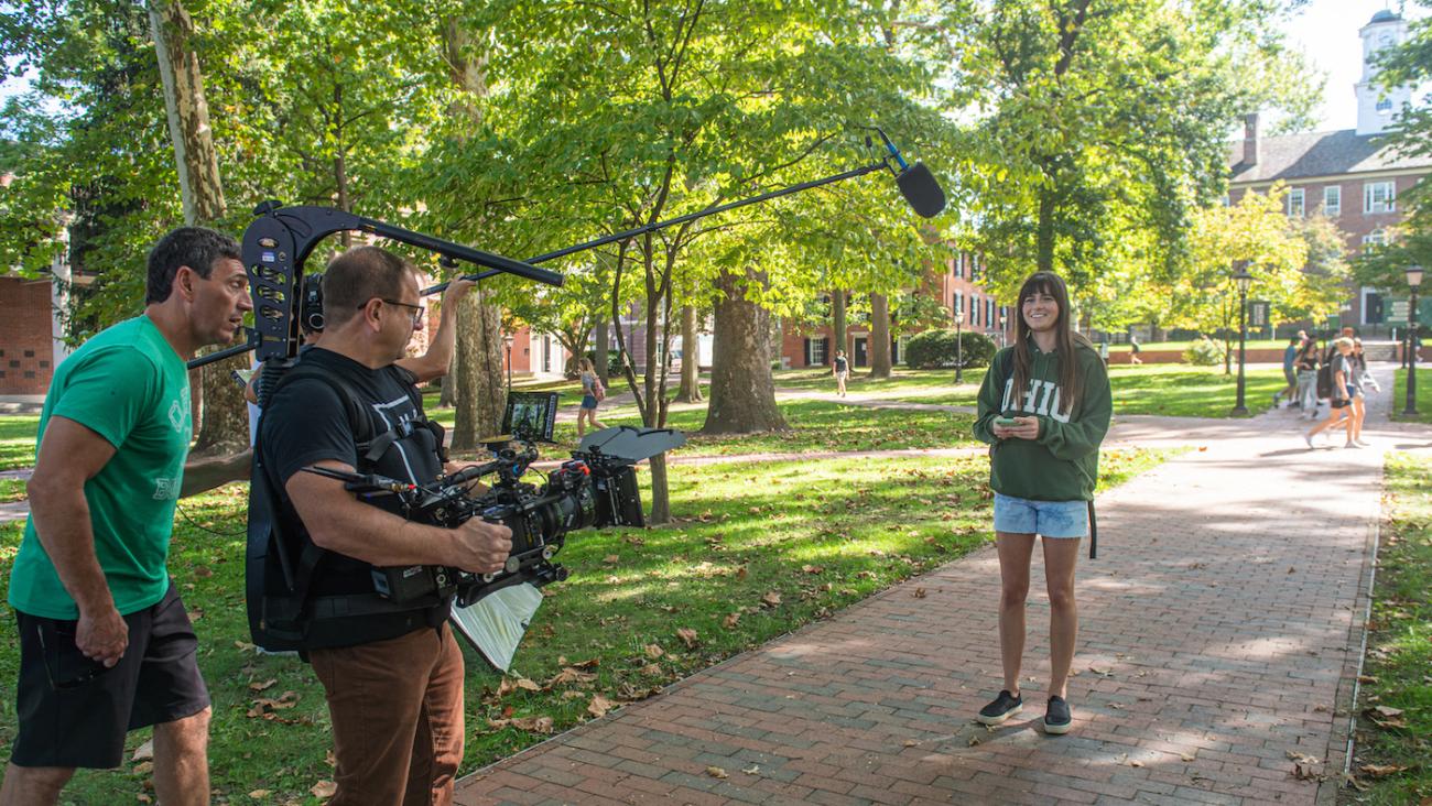 A man is holding a film camera pointed towards a student on College Green