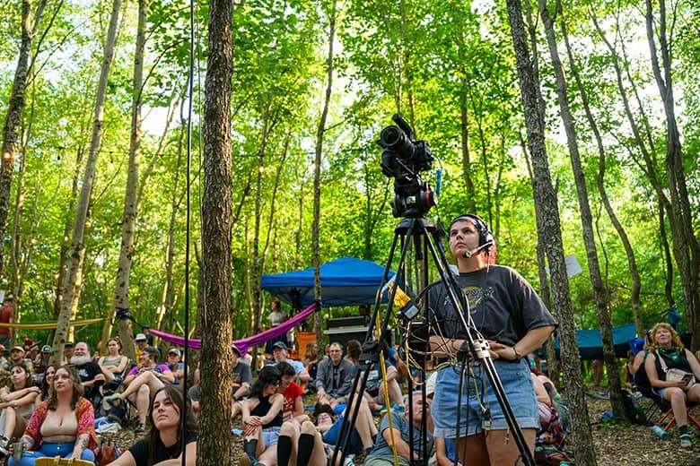 An OHIO student stands behind a camera with a tripod and films the Creekside Stage with the audience in the background.