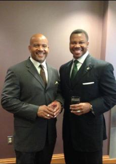 two men pose smiling in dark suits