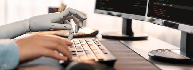 A robot hand typing on computer keyboard next to a human hand.