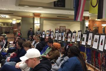 The crowd at the Trans* Day of Remembrance ceremony sits in front of posters of victims of violence in the U.S.