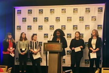 Six students stand on stage, with one at the podium while presenting