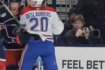 Two hockey players collide in the foreground as a photographer observes from the stands