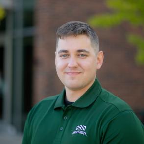 Brian Wolfe stands in front of a brick building, smiling at the camera, wearing a dark green 'Ohio University' shirt.