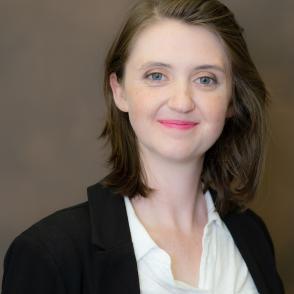 Liz Myers stands in front of a brown background, wearing a white shirt and black blazer, giving the camera a half-smile.