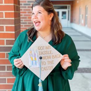 Brooke Kelley wears a green, long-sleeved dress. Brooke looks away from the camera with a grin, holding out a graduation cap that reads 'got my tassel, now back to the castle.'