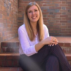 Jane Dreher sits on brick stairs and smiles at the camera, wearing a long sleeve pale pink button-up and navy and dark red pinstripe pants.