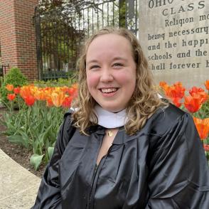 Lauren Gaines sits in front of the Ohio University entrance in a graduation gown and hood. Lauren smiles at the camera.
