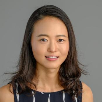 Headshot of Zeyu Zhang. Woman smiling at camera with shoulder length brown hair wearing a navy and white striped shirt. Her background is a neutral middle gray. 